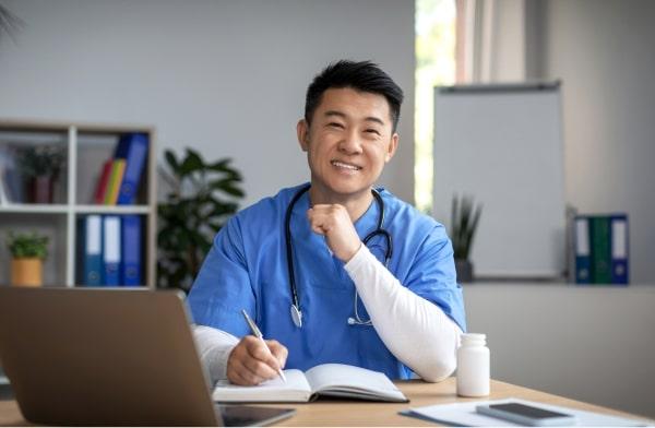 /cheerful-young-korean-male-doctor-uniform-with-stethoscope-workplace-work-with-laptop.jpg
